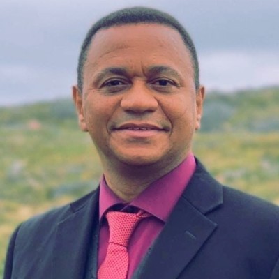 Black man with short hair, in an image taken outside in front of a hill. He is wearing a maroon dress shirt, a pink tie, and a black suit jacket.